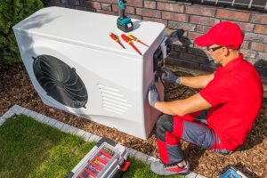 Professional Middle Aged HVAC Technician in Red Uniform Repairing Modern Heat Pump Unit. House Heating and Cooling System Theme.